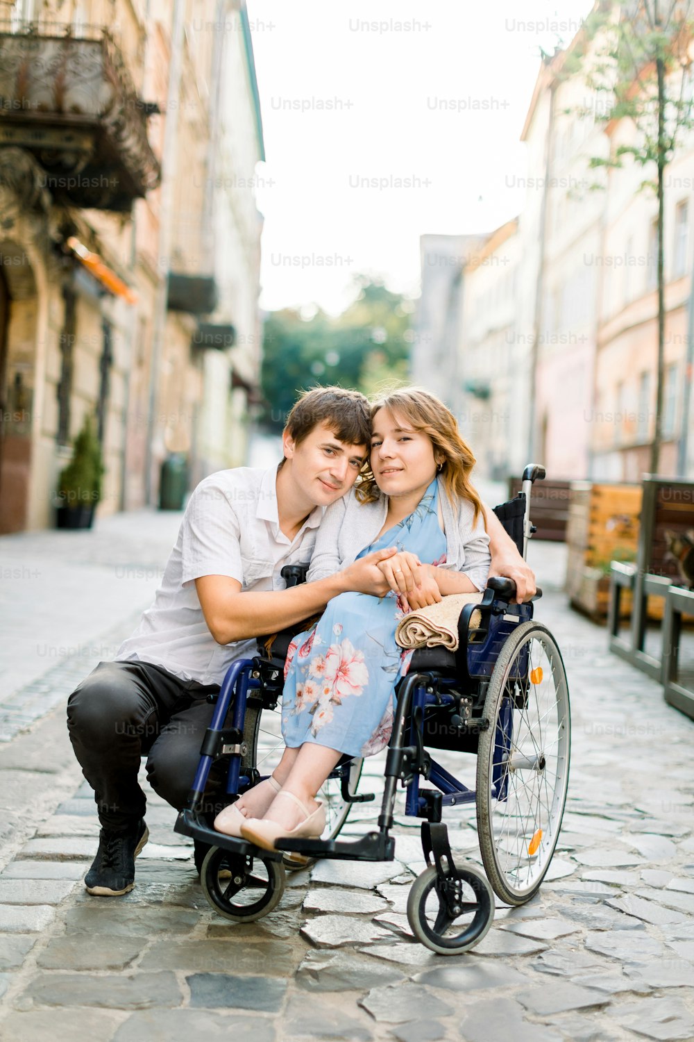 Mulher sorridente muito jovem na cadeira de rodas e homem bonito olhando para a câmera, de mãos dadas, apaixonado, andando no centro da cidade velha