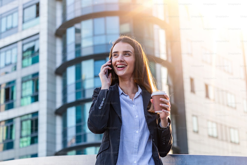 Business woman with coffee and talking on the phone near office. Young woman with smartphone standing against street blurred building background. Beautiful girl in casual suite with phone and coffee