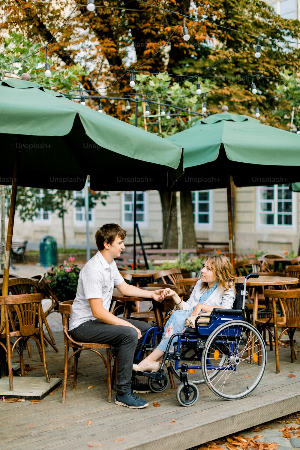휠체어를 탄 젊은 예쁜 여자가 남자 남자친구와 손을 잡고 구시가지의 야외 카페에서 시간을 보내고 있다.