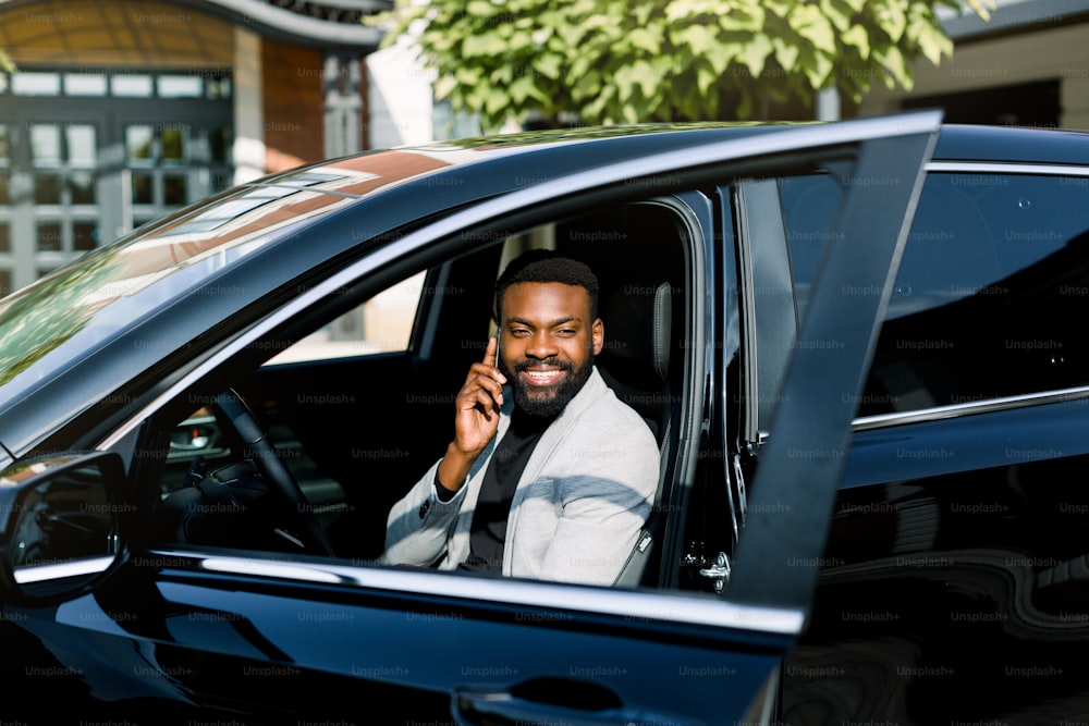 Hombre africano alegre en chaqueta gey sentado en el coche hablando por teléfono, centro de oficina en el fondo.