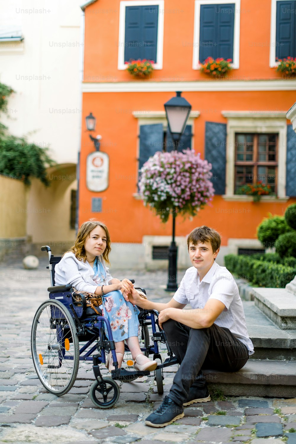 Couple in love in old city center. Pretty young girl with disease on a wheelchair and her lovely man, sitting on the stairs. Stylish orange ancient building on the background.