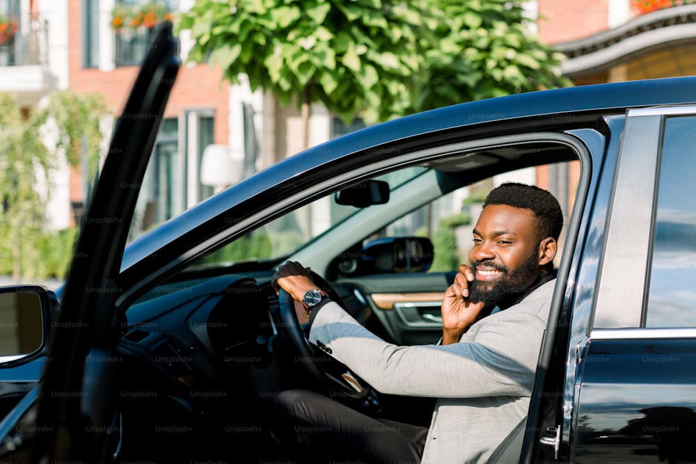 young African businessman sitting in his black car and making a phone call, and smiling. Office center buildings on the background.