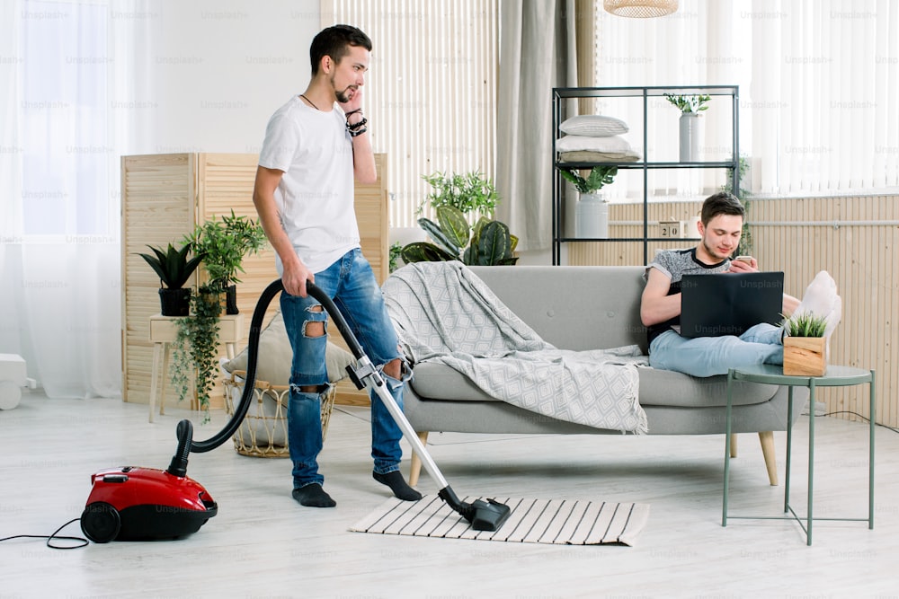 Couple of gay cleaning living room at home, Young gay man holding the laptop, sitting sofa. Young gay with apron using vacuum holiday together