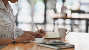 Cropped shot of young secretary woman in white striped shirt sitting at the wooden desk and writing on the notebook/taking notes.