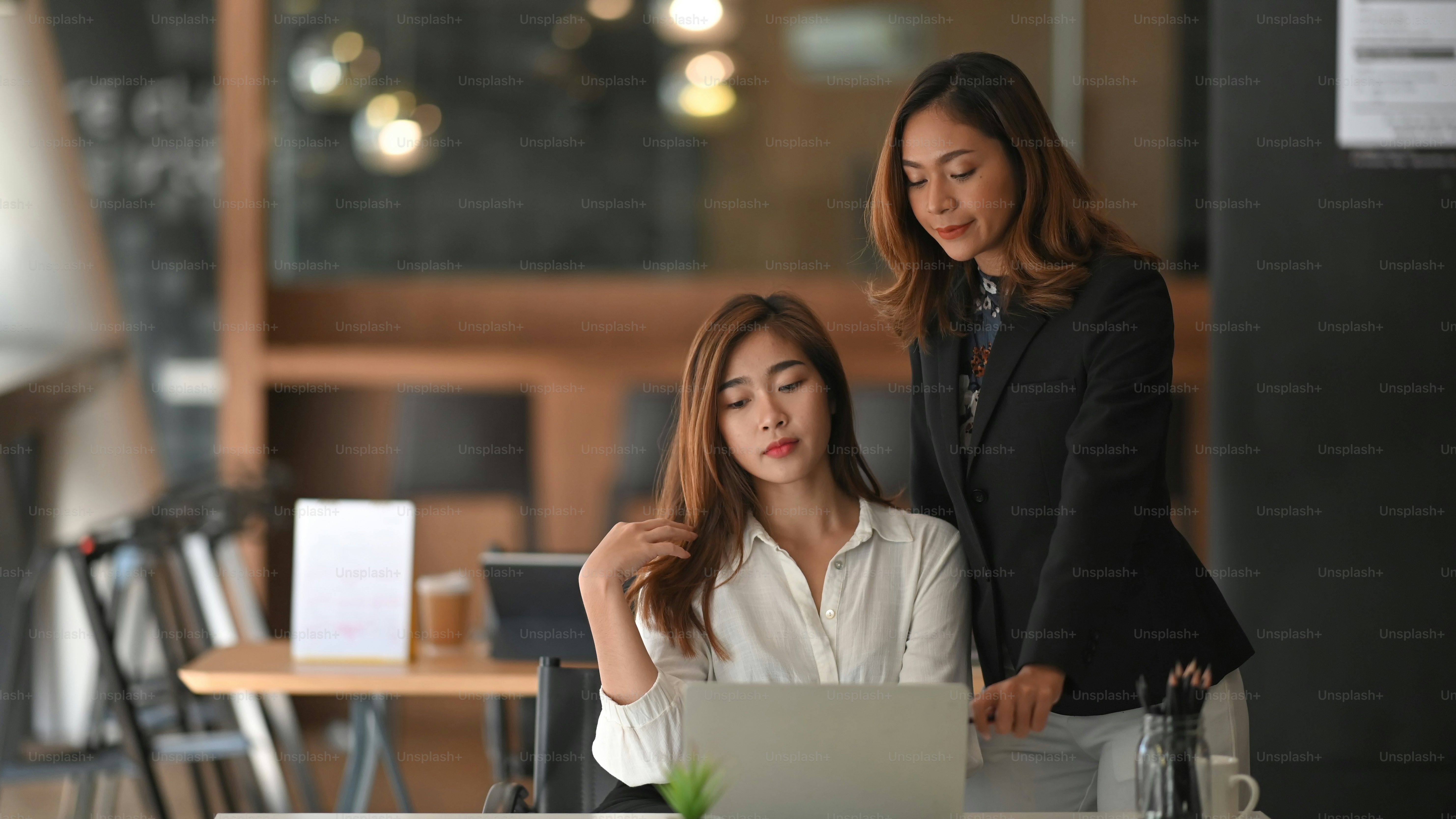 Confident business woman in black suit standing and advising to beautiful white shirt employee that sitting/posing over the modern office background.