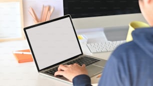 Detrás de la toma de un programador inteligente que sostiene una taza de café y escribe en una computadora portátil con pantalla blanca en blanco mientras está sentado sobre su monitor de pantalla negra en un escritorio de trabajo moderno.