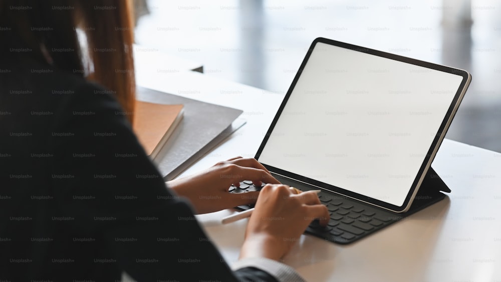 Atrás de uma foto de uma jovem empresária digitando no teclado do tablet com tela branca em branco enquanto estava sentada na moderna mesa de trabalho.