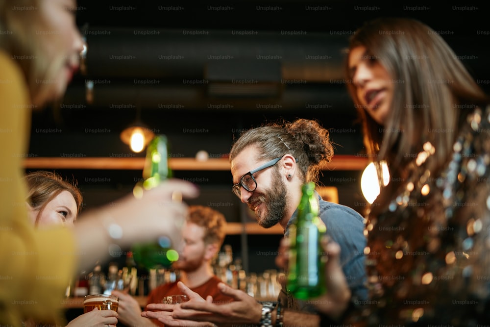 Um pequeno grupo de melhores amigos em pé em um pub, bebendo cerveja, conversando e se divertindo. Concentre-se no barman limpando o copo. Vida noturna.