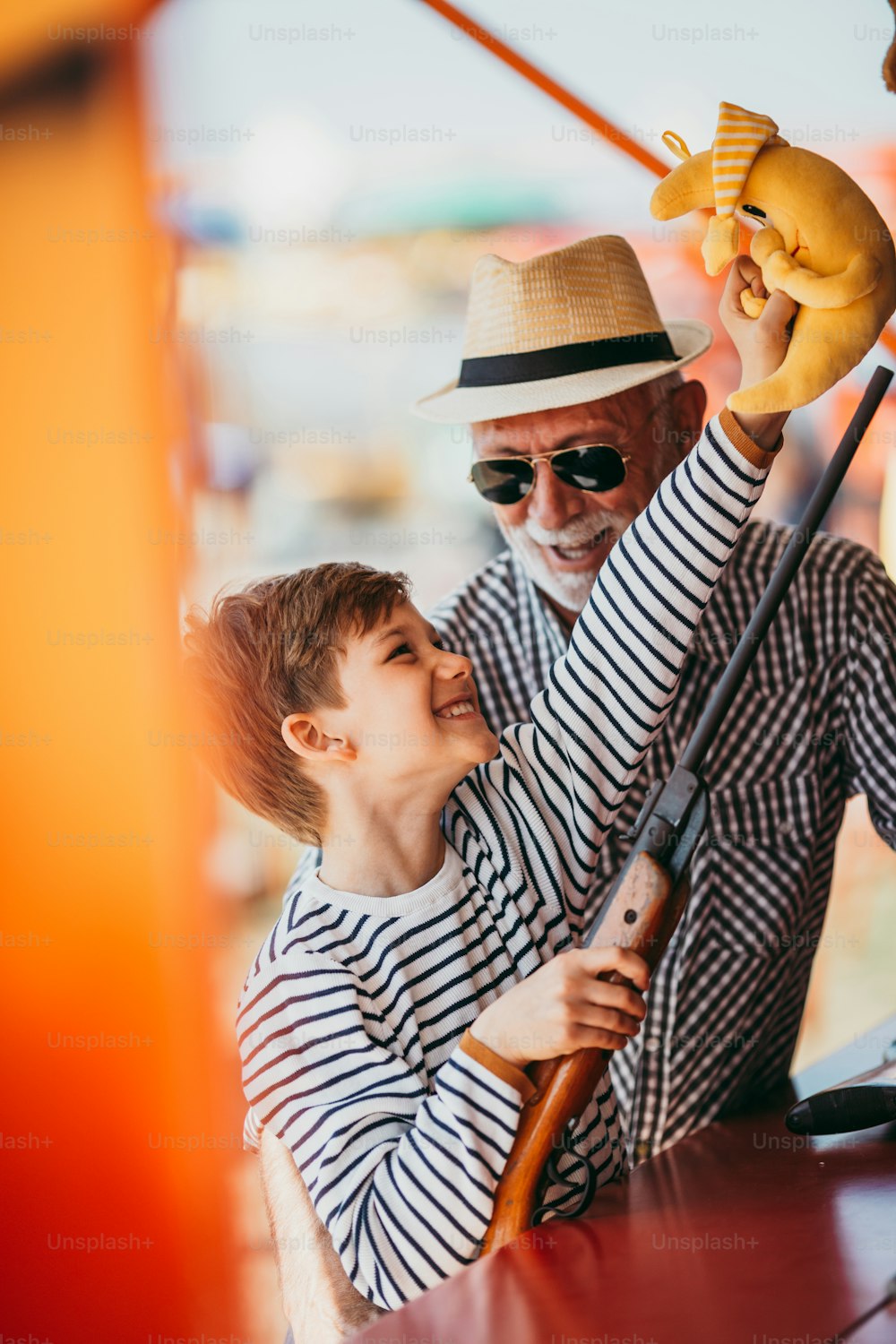 Abuelo y nieto divirtiéndose y pasando tiempo de buena calidad juntos en el parque de atracciones. Niño disparando con pistola de aire comprimido mientras el abuelo lo ayuda a ganar el premio.