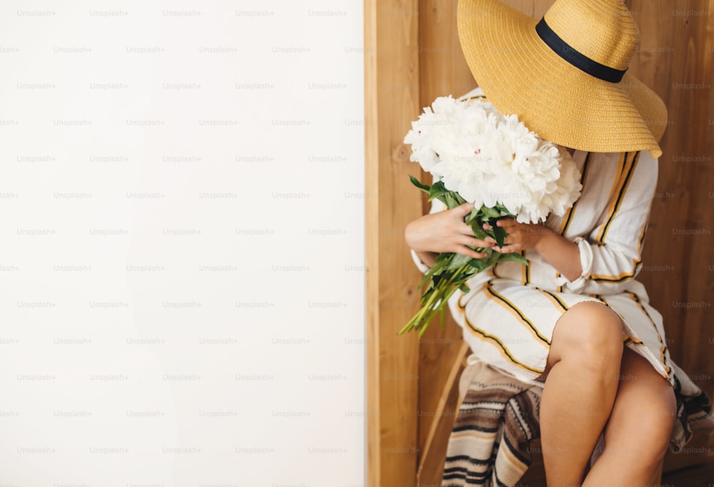 Stylish boho woman in hat holding white peony bouquet on rustic wooden background. Space for text. Hipster girl in dress posing with peonies. Happy Mothers day. International Womens days
