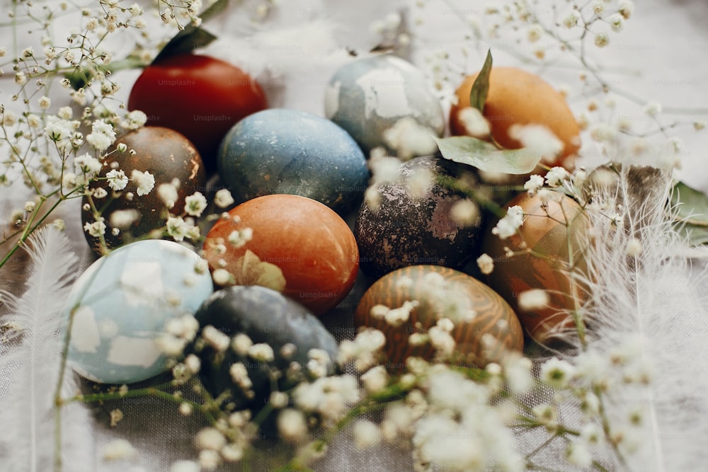 Oeufs de Pâques élégants dans un nid rustique sur la table. Oeufs de Pâques colorés teints au naturel avec des fleurs blanches de printemps et des plumes sur fond textile rural. Joyeuses Pâques