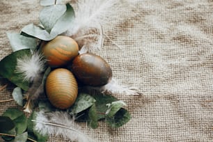 Stylish easter eggs in rustic nest with feathers on rustic table. Natural dyed green easter eggs with eucalyptus branch, spring flowers on rural textile background. Flat lay