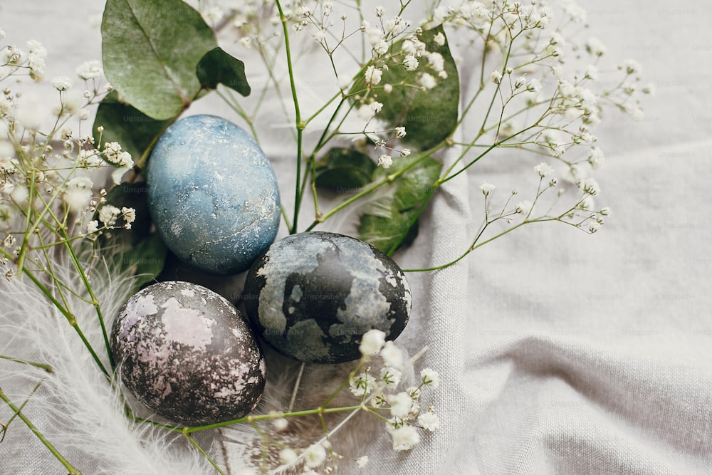 행복한 부활절. 테이블에 소박한 둥지에서 세련된 부활절 달걀. 시골 섬유 배경에 봄 흰색 꽃과 깃털이 있는 자연 염색 다채로운 부활절 달걀. 복사 공간