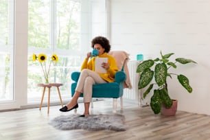 Beautiful woman sitting in an armchair, reading an e-book on a tablet computer and drinking tea; woman enjoying her leisure time activities at home