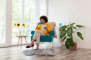 Bella donna seduta su una poltrona, che legge un e-book su un tablet e beve tè; donna che si gode le sue attività del tempo libero a casa
