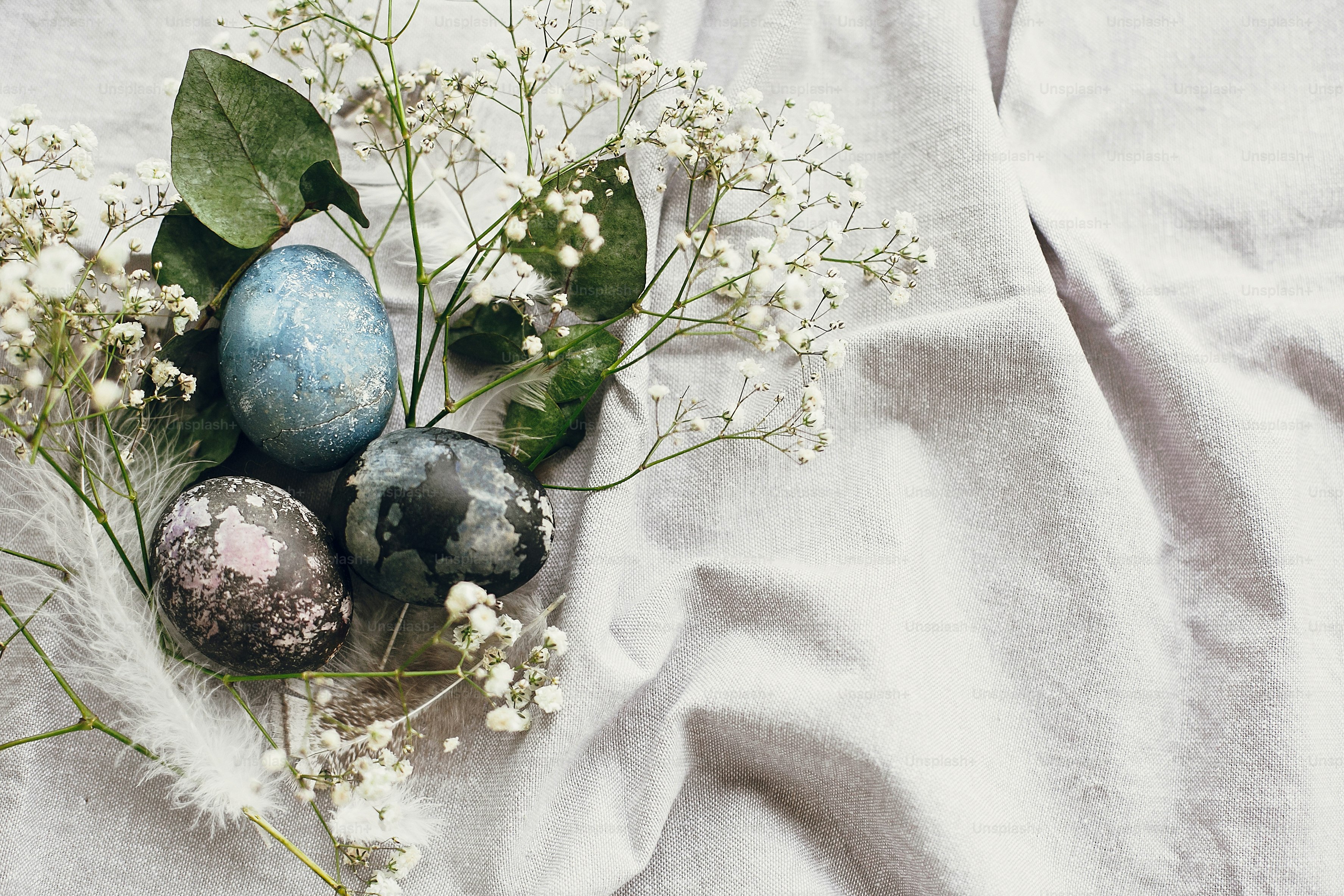 Stylish easter eggs in rustic nest on table flat lay. Natural dyed colorful easter eggs with spring white flowers and feathers on rural textile background. Copy space