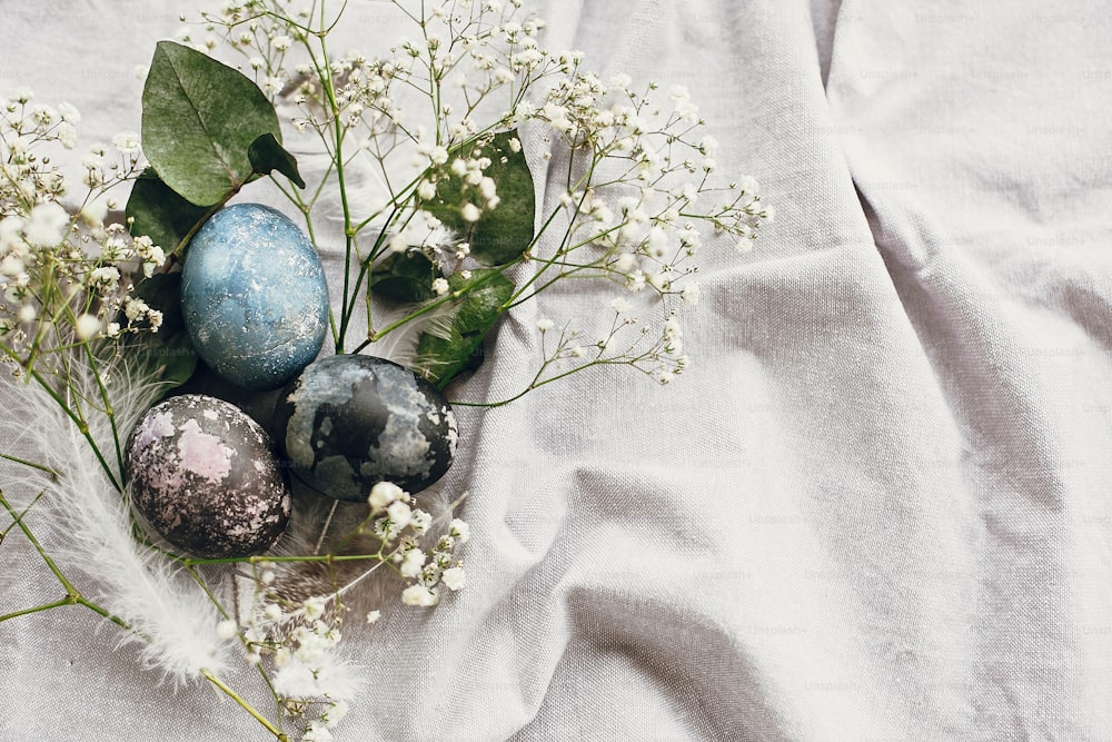 Stylish easter eggs in rustic nest on table flat lay. Natural dyed colorful easter eggs with spring white flowers and feathers on rural textile background. Copy space