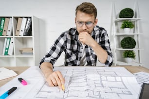 Minder und intelligenter junger Erwachsener Architekt, der mit Blaupausen im Arbeitsplatzbüro arbeitet, die Lösung für die Innenarchitektur in einem neuen Haus prüft, ein nachdenkliches Gesicht macht