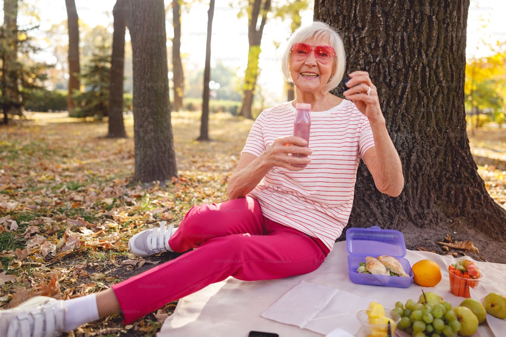 Gut aussehende alte Frau, die im Park auf dem Boden sitzt und Smoothie isst
