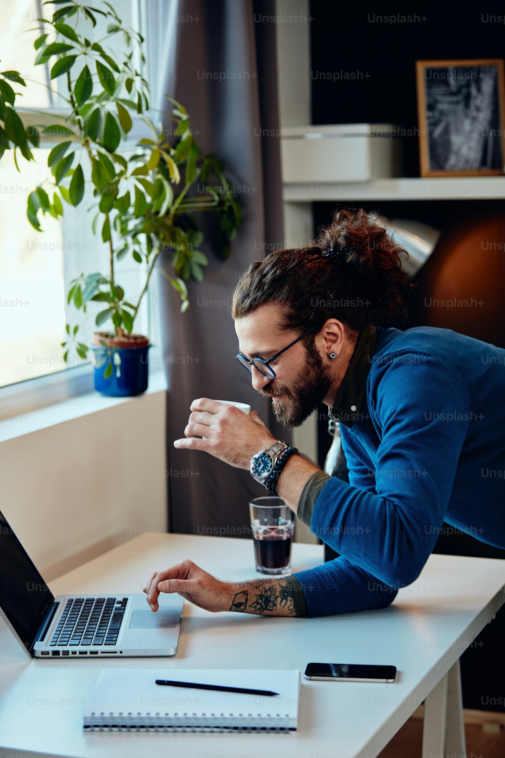 Joven hipster barbudo atractivo con cabello rizado apoyado en la mesa, bebiendo café y navegando por Internet mientras está de pie en la oficina en casa.