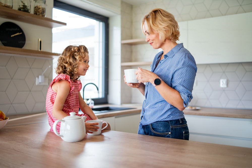 A happy cute small girl with mother indoors in kitchen at home, drinking tea.