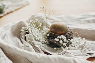Stilvolles Osterei im rustikalen Nest auf Holztisch. Natürlich gefärbtes grünes Osterei mit weißen Frühlingsblumen auf ländlichem Textilhintergrund. Platz für Text