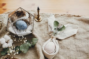 Stylish easter eggs, modern nest, napkin bunny ears on rustic table. Natural dyed easter eggs with eucalyptus branch, spring flowers, cotton on rural background. Table setting