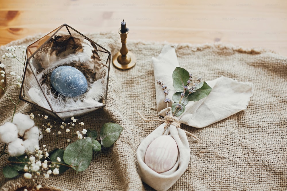 Stylish easter eggs, modern nest, napkin bunny ears on rustic table. Natural dyed easter eggs with eucalyptus branch, spring flowers, cotton on rural background. Table setting