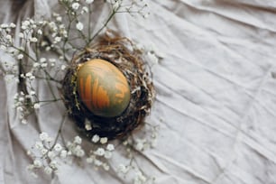 Stilvolles Osterei im rustikalen Nest auf Holztisch. Natürlich gefärbtes grünes Osterei mit weißen Frühlingsblumen auf ländlichem Textilhintergrund. Platz für Text. Flache Liege