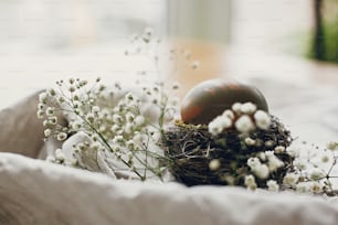 Stilvolles Osterei im rustikalen Nest auf Holztisch. Natürlich gefärbtes grünes Osterei mit weißen Frühlingsblumen auf ländlichem Textilhintergrund. Platz für Text