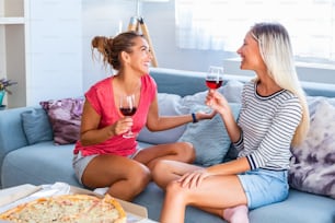 Two attractive girls, cheerful best friends having fun and drinking red wine at home. Two glasses of white wine in hands. Girls having fun time together gossiping