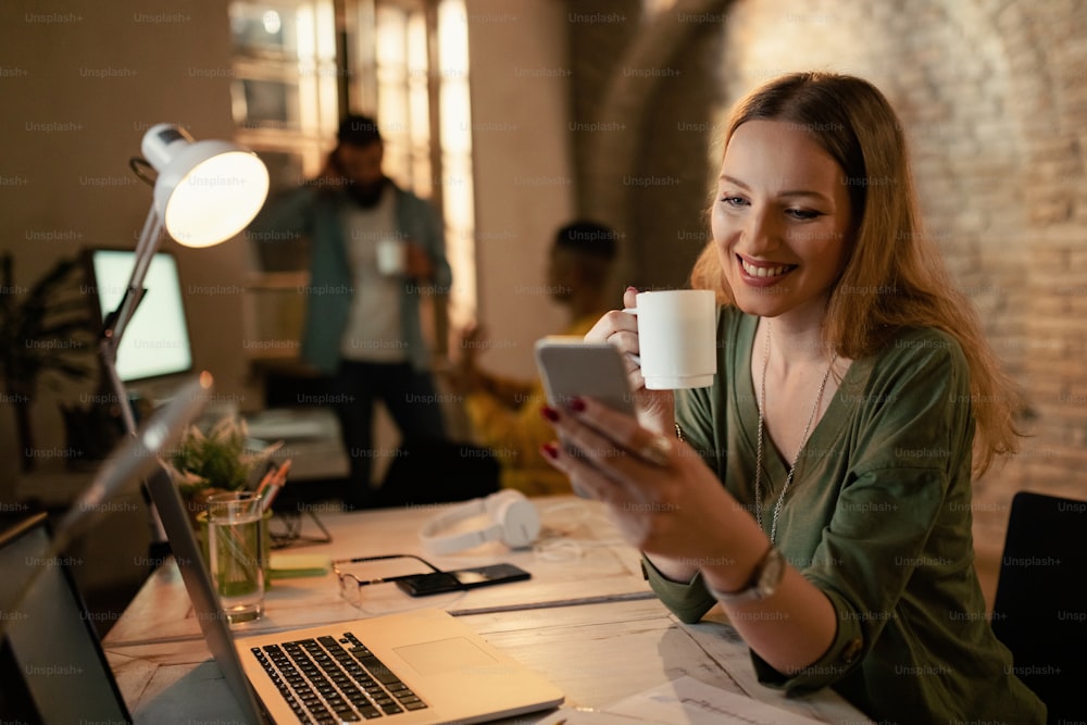 Glückliche Unternehmerin, die SMS auf dem Smartphone liest, während sie nachts im Büro Kaffee trinkt.