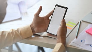 Close-up image of young creative man is holding crop black smartphone with white blank screen on working desk.