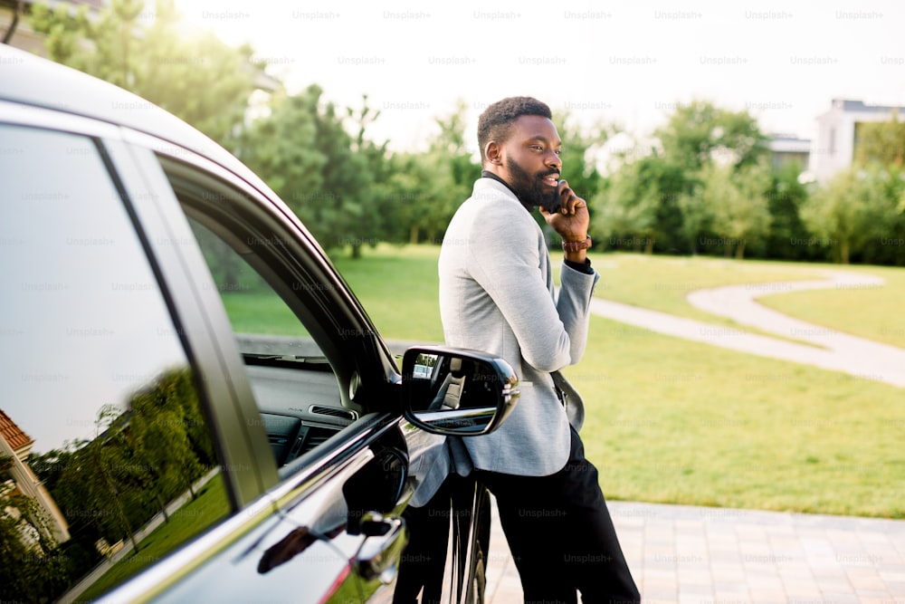Un hombre de negocios afroamericano con traje habla por teléfono inteligente mientras está parado al aire libre cerca de su lujoso automóvil negro. Parque verde, árboles al fondo.