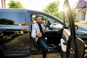 Successful handsome African man businessman and boss sitting in the car, talking on the phone, smiling, looking at camera and showing thumb up. Business concept.