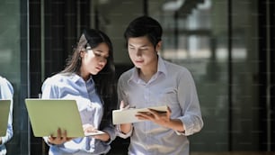 Foto de um jovem casal de startups de negócios em pé juntos segurando / olhando para laptop e tablet enquanto conversam / discutem / fazem brainstorming sobre o processo de negócios deles.