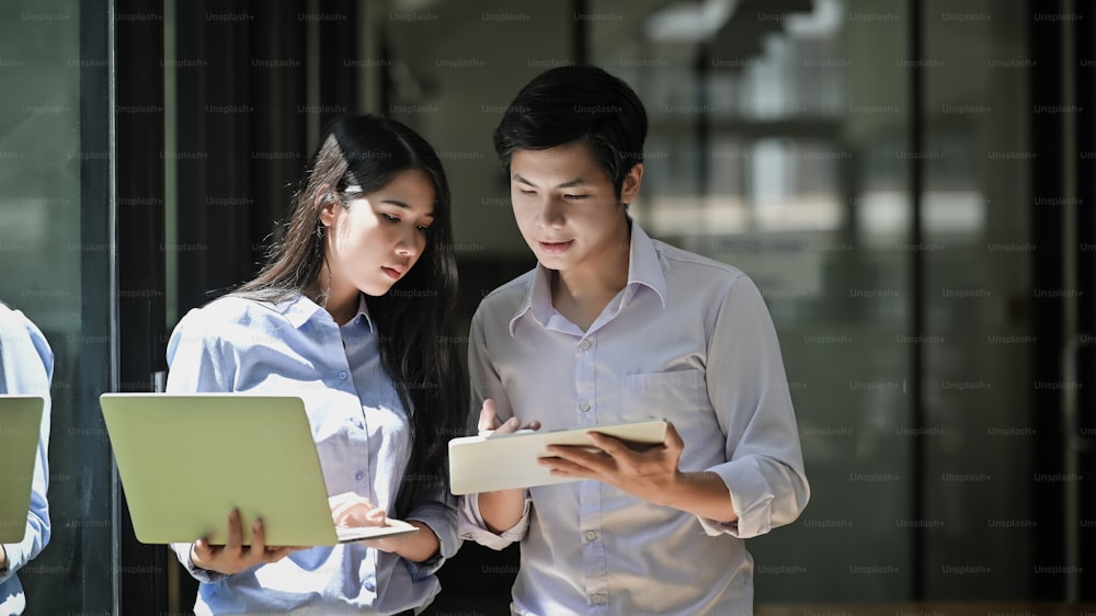Photo of young business startup couple standing together holding/looking at laptop and tablet while talking/discussing/brainstorming about them business process.