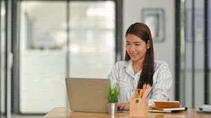 Business girl in striped shirt typing on laptop and sitting at the wooden working desk with modern office background.