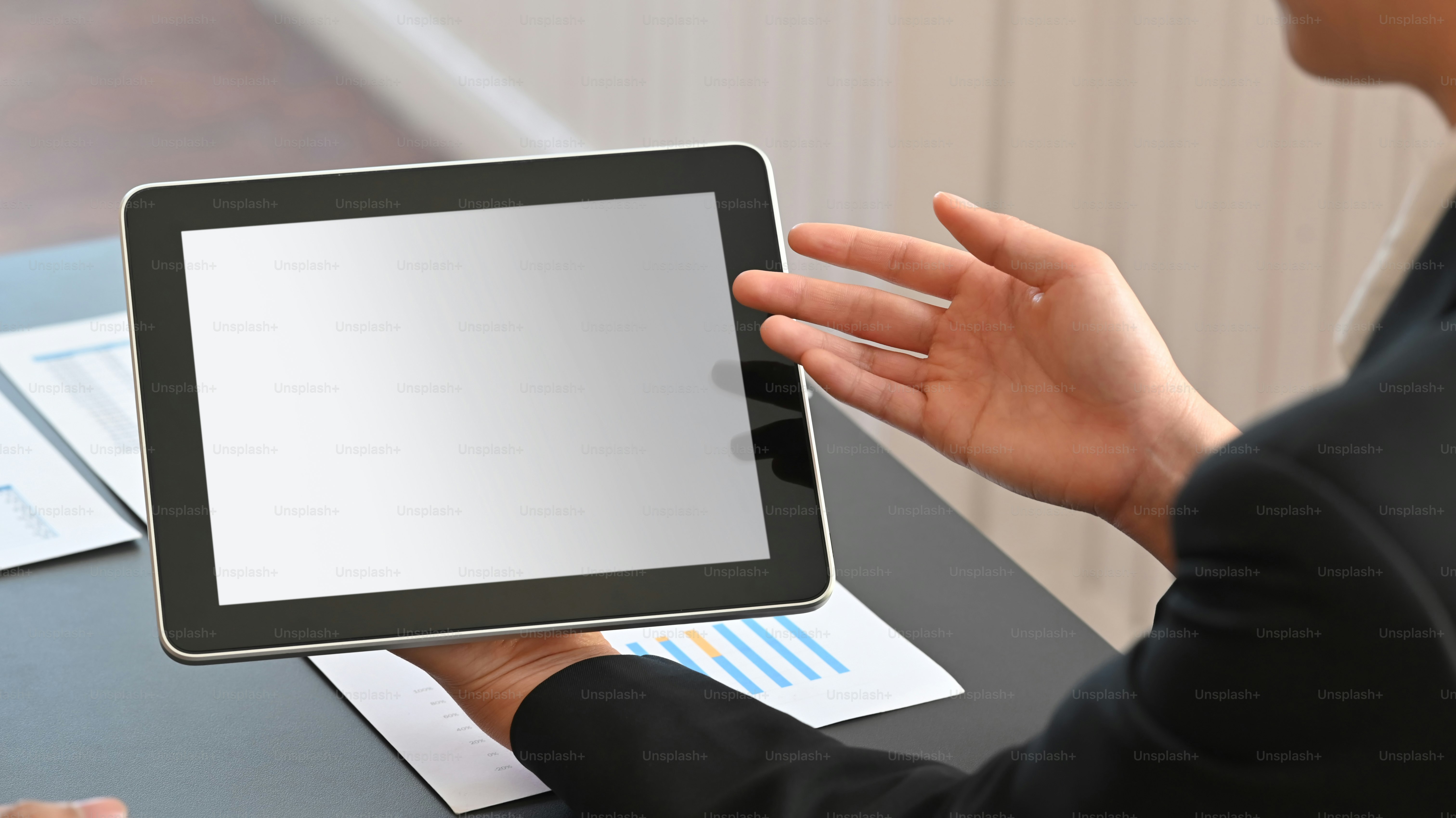 Cropped shot of smart businessman in black suit holding crop black tablet with white blank screen in hand while sitting at the modern table with modern meeting room as background.