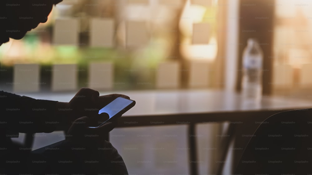 Imagen de la silueta del hombre sentado en el escritorio de trabajo moderno y navegando por la red por su teléfono inteligente con la puesta de sol borrosa como fondo.