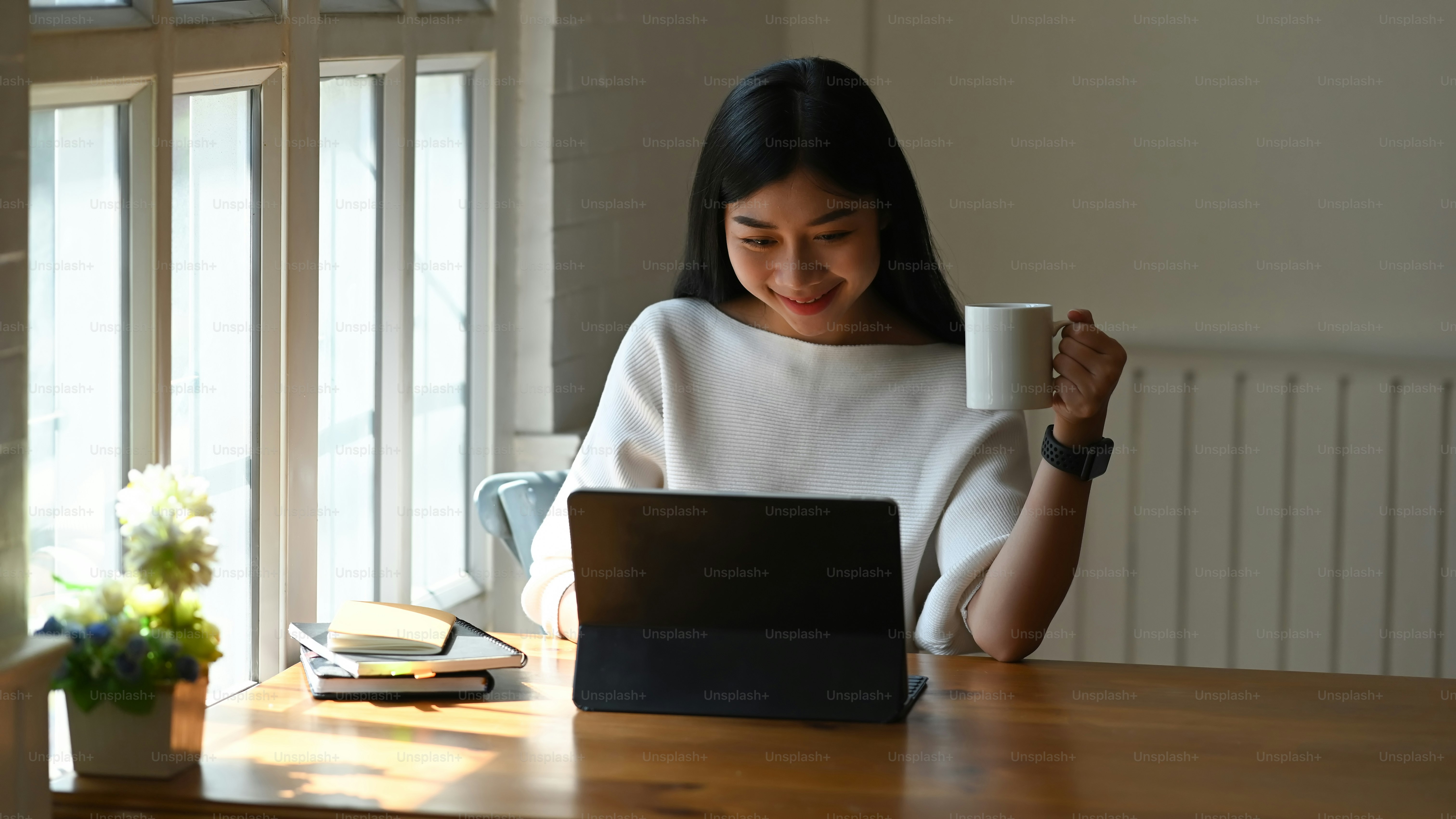 Attractive woman working her tablet computer and holding coffee mug in home office.
