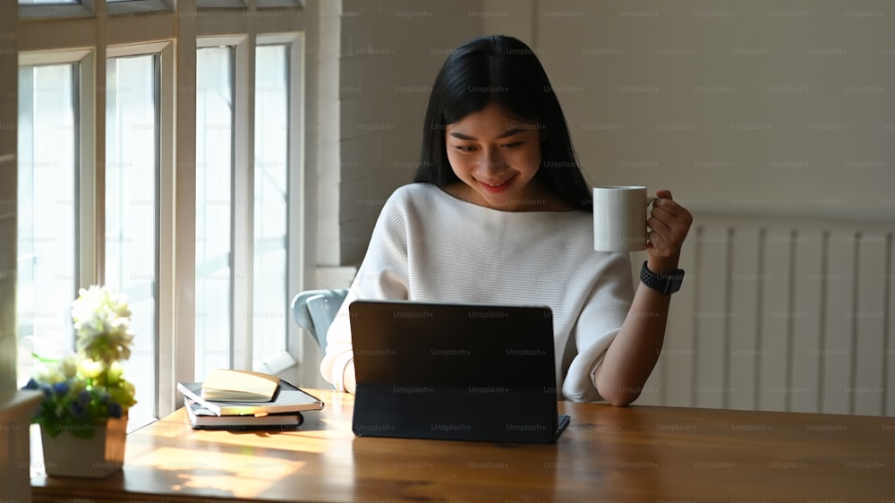 Attractive woman working her tablet computer and holding coffee mug in home office.