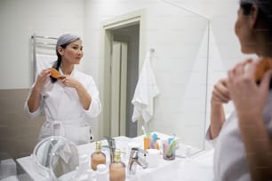 Peaceful adult woman in bathrobe looking at her reflection in the mirror and brushing hair