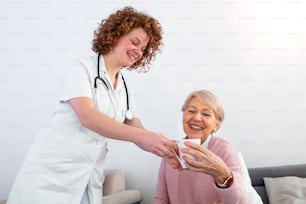 Pretty young caregiver serving afternoon cup of tea to older happy woman. Young nurse caring for elderly patient in her home. Dementia and Occupational Therapy Home caregiver and senior adult woman.