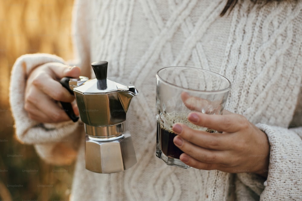 Donna hipster che tiene la caffettiera geyser e la tazza di vetro con caffè in luce calda e soleggiata nelle erbe della campagna rurale. Momento rustico atmosferico. Preparazione alternativa del caffè in viaggio.