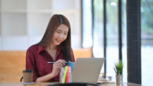 photo of young beautiful designer in red shirt taking notes in front of her laptop at the wooden desk over the modern office background.