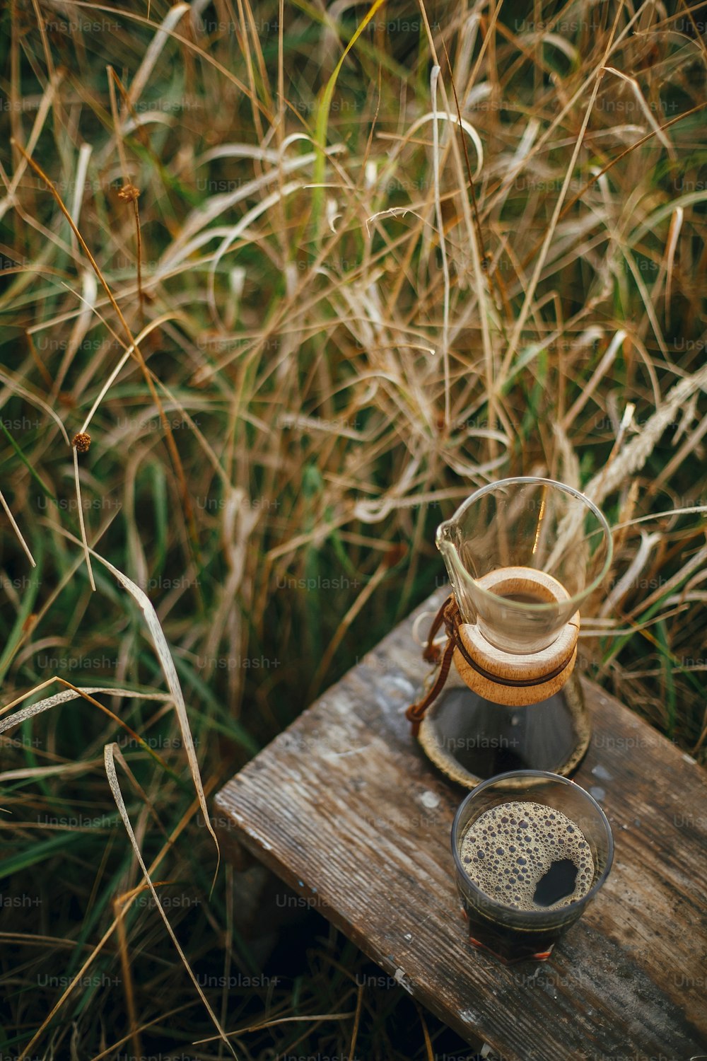 Café caliente en taza de vidrio y frasco de vidrio en fondo en luz cálida soleada en hierbas rurales. Preparación alternativa de café al aire libre en viajes. Momento tranquilo rústico atmosférico. Imagen vertical