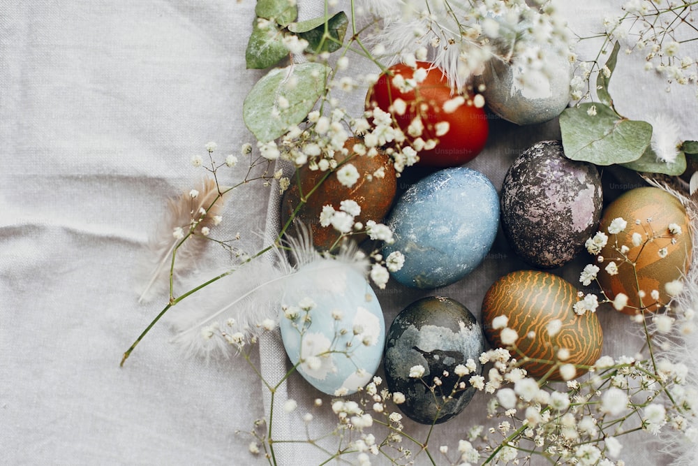 테이블 플랫 레이에 소박한 둥지에 세련된 부활절 달걀. 시골 섬유 배경에 봄 흰색 꽃과 깃털이 있는 자연 염색 다채로운 부활절 달걀. 행복한 부활절