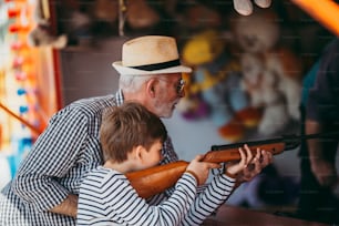 Abuelo y nieto divirtiéndose y pasando tiempo de buena calidad juntos en el parque de atracciones. Niño disparando con pistola de aire comprimido mientras el abuelo lo ayuda a ganar el premio.