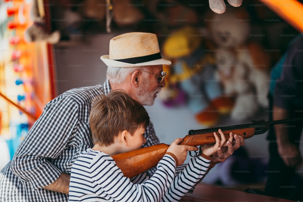Abuelo y nieto divirtiéndose y pasando tiempo de buena calidad juntos en el parque de atracciones. Niño disparando con pistola de aire comprimido mientras el abuelo lo ayuda a ganar el premio.
